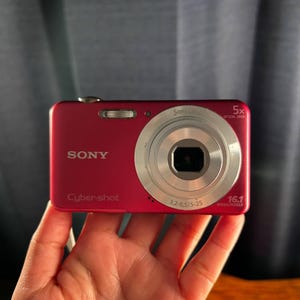 Puede incluir: Una cámara digital Sony Cyber-shot de color magenta con un barril de lente plateado, zoom óptico de 5x y un sensor de 16,1 megapíxeles. La parte frontal muestra el logotipo de Sony, la marca Cyber-shot y las especificaciones 3,2-6,5/5-25.