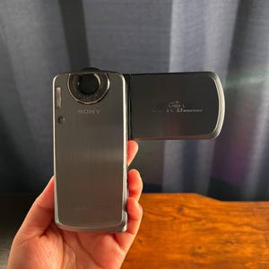 May include: A silver Sony Cyber-shot digital camera with a rotating lens and a flip-out screen. The camera body features the Sony logo and the Cyber-shot branding. The screen displays text including 5.1 Megapixels and Stereo L.