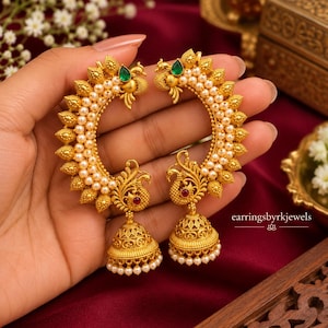 May include: A pair of gold-toned crescent-shaped earrings featuring intricate peacock motifs, small white pearls, and a single green teardrop stone at the top. Each earring includes a dangling bell-shaped jhumka with a small red stone and pearls.