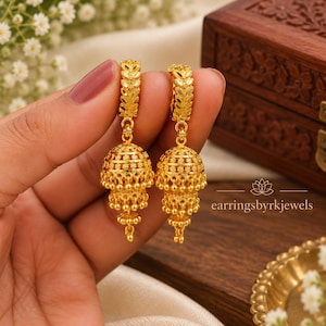 Könnte beinhalten: Ein Paar goldene Jhumka-Ohrringe mit einem strukturierten, blattförmigen Creolen-Oberteil und einem mehrstufigen, glockenförmigen Anhänger mit filigranen Details und kleinen hängenden Perlen. Der Text earringsbyrkjewels ist rechts zu sehen.