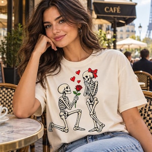 May include: A cream-coloured short-sleeve t-shirt featuring a graphic print of two skeletons. One skeleton is kneeling and offering a red rose to the other skeleton, who wears a red bow. Three small red hearts float above the skeletons.