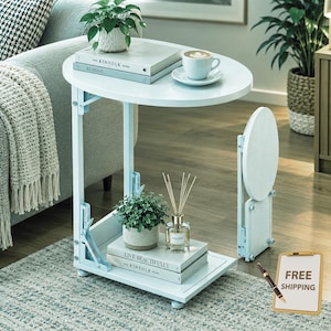 May include: A white C-shaped side table with an oval top and a rectangular base. The table features a folding mechanism, a lower shelf holding a potted plant and a glass reed diffuser, and an upper surface with a book and a coffee cup.