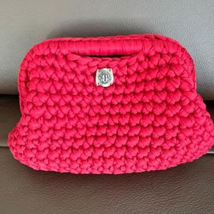 May include: A vibrant red crochet clutch bag featuring a thick, textured yarn weave and a central silver-toned metal emblem with the letter T engraved inside a decorative circular border, positioned on the front flap of the handbag.