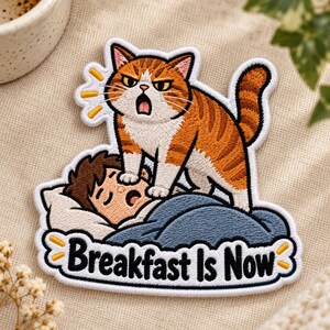 May include: An embroidered patch featuring an orange and white tabby cat standing on a person sleeping in a bed under a blue blanket. The cat has an open mouth and yellow sound lines. The text "Breakfast Is Now" is printed in black at the bottom.
