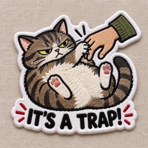 May include: An embroidered patch featuring a grumpy brown tabby cat lying on its back, swatting at a hand wearing a green sleeve. The cat has white fur on its belly and paws with pink pads. The text IT'S A TRAP! is embroidered in black below.
