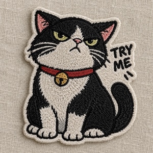 May include: An embroidered patch featuring a black and white cat with a grumpy expression, wearing a red collar with a gold bell. The cat is sitting, and the text TRY ME is embroidered in black to the right of the cat's head.