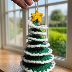 May include: A spiral-shaped Christmas tree ornament crafted from green yarn with white trim along the edges. The tree features a bright yellow crocheted star at the top and is suspended by a thin, light brown twine loop for hanging purposes.