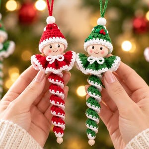 May include: Two crochet ornaments shaped like figures with round faces, wearing pointed hats and ruffled collars. One figure is red and white, the other is green and white. Both have spiral-shaped bodies and are suspended by thin strings.