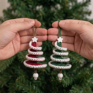 Pode incluir: Dois enfeites de árvore de Natal em forma de espiral feitos de fio de crochê. Uma árvore é vermelho-escura com acabamento branco e a outra é verde-escura com acabamento branco. Cada uma tem uma pequena estrela branca e uma conta branca.