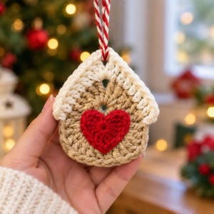 May include: A house-shaped crochet ornament featuring a tan base, a cream-colored roof, and a bright red heart stitched in the center. The ornament is suspended by a red and white twisted string loop for hanging on a holiday tree.