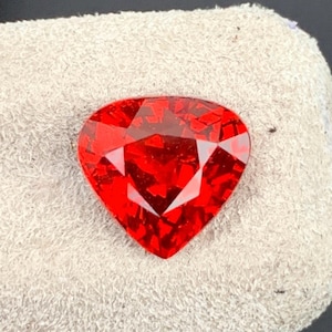 Puede incluir: Una gema de corte triangular en un tono rojo vibrante con una superficie facetada que crea un efecto reflectante y brillante. La piedra presenta bordes geométricos definidos y un acabado pulido que muestra profundos tonos carmesí.