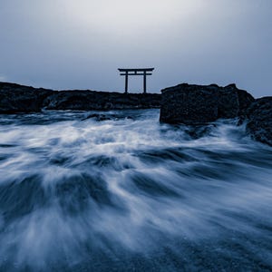 以下が含まれることがあります： 暗い青色の色調の風景で、岩の多い海岸線に伝統的な鳥居が立っています。前景には、薄暗い夕暮れの空の下、暗くギザギザした岩に打ち寄せる、動きを捉えたぼやけた海の波が写っています。