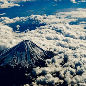 以下が含まれることがあります： 雲海の上にそびえ立つ富士山の航空写真。山頂付近は雪で白く覆われ、山肌は暗い岩肌が露出している。青い空を背景に、白い雲が山を取り囲むように広がっており、円錐形の火山特有の山容がはっきりと捉えられている。