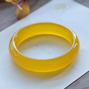 Op de afbeelding: Een gladde, doorschijnende gele armband met een afgerond profiel. Het cirkelvormige sieraad heeft een gepolijste, glanzende afwerking die het licht opvangt, waardoor subtiele accenten langs het gebogen oppervlak en een heldere uitstraling ontstaan.