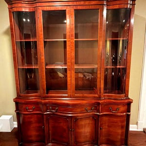May include: A large, reddish-brown wooden china cabinet featuring four glass-fronted upper doors with internal shelving, and a lower section with four drawers and four solid cabinet doors, all accented with curved lines and metal hardware.