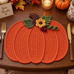 May include: A pumpkin-shaped crochet placemat in vibrant orange with a dark brown stem, two green leaves, a yellow flower with a brown center, and two small dark red berries attached to the top, creating a textured, handcrafted table accessory.