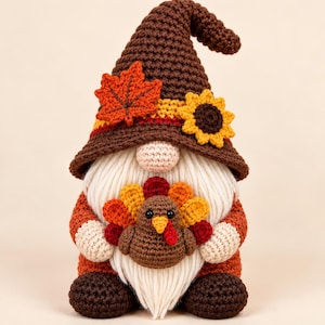May include: A crochet gnome figurine featuring a tall brown pointed hat decorated with a crocheted orange maple leaf and a yellow sunflower. The gnome has a cream-colored beard, a round nose, and holds a small brown turkey with colorful tail feathers.