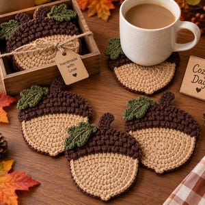 May include: A set of crochet acorn-shaped coasters in tan and dark brown with green leaves. Some coasters are stacked in a wooden box with a tag reading Handmade with Love, while others are placed on a surface with a white mug and a sign reading Cozy Days.