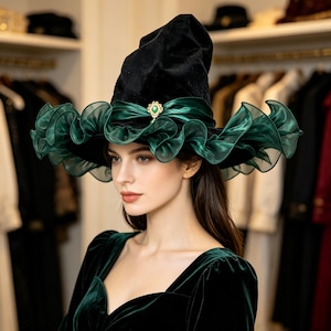 Peut inclure: Un chapeau pointu en velours noir doté d'un large bord à volants en organza vert foncé. Un ruban de satin vert foncé entoure la base de la calotte, fixé à l'avant par une broche dorée ornée d'une pierre précieuse ovale de couleur verte.