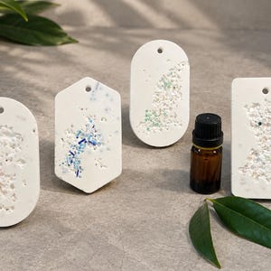 May include: Four white hanging ornaments in oval, hexagonal, and rectangular shapes with textured, porous surfaces containing small colourful flecks. Each piece features a small hole at the top. A small amber glass bottle with a black cap is included.