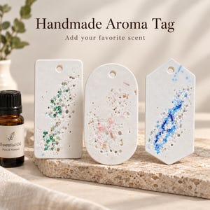 May include: Three white aroma tags in rectangular, oval, and hexagonal shapes with textured surfaces containing embedded green, pink, and blue beads. Each tag features a small hole at the top. A brown glass bottle labeled Essential Oil sits nearby.