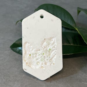 May include: A hexagonal off-white hanging ornament with a smooth finish and a small circular hole at the top. The center features a textured, recessed area containing small, translucent, light green circular beads embedded in the surface.