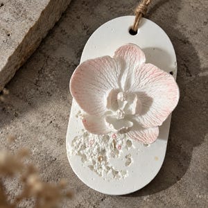Puede incluir: Un adorno colgante ovalado de color blanco con una flor de orquídea tridimensional de color rosa pálido y blanco fijada en el centro. La parte inferior del adorno tiene una superficie texturizada y porosa, y lleva un lazo de cuerda marrón en la parte superior.