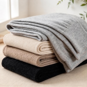 May include: A stack of four folded knit garments in neutral tones. The top piece is light gray, followed by a cream-colored item, a tan piece, and a black garment at the bottom. Each piece features a soft, textured knit fabric.