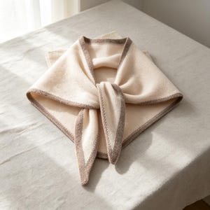 Peut inclure: Un foulard triangulaire de couleur crème douce avec une bordure contrastante beige foncé sur tous les bords. Le foulard est plié et noué au centre, mettant en valeur une texture tricotée sur tout le tissu et une finition soignée.
