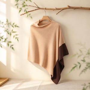 May include: A two-toned poncho featuring a large beige section and a smaller dark brown section at the corner. The garment is made of a soft knit fabric and is displayed hanging from a wooden hanger on a branch against a plain wall.