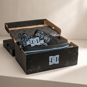 May include: A black sneaker with a light blue DC logo and intricate paisley-style patterns in light blue and white. The shoe features black laces and a light blue sole. It is presented inside a black cardboard box with the DC logo printed on the front.