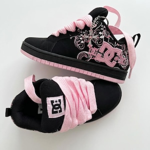 Op de afbeelding: Een paar zwarte en roze DC-sneakers met dikke roze veters, een roze DC-logo aan de zijkant en een zilveren kruisbedel bevestigd aan de oogjes. De schoenen hebben een zwarte basis met roze bloemmotieven en een roze rubberen zool.