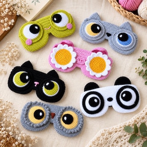 Könnte beinhalten: Sechs gehäkelte Schlafmasken mit verschiedenen Tier- und Blumenmotiven. Die Kollektion umfasst einen lindgrünen Frosch, eine graue Katze, eine rosa Gänseblümchenblüte, eine schwarze Katze, einen weißen Panda und eine graue Eule aus Garn.