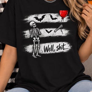 May include: A black t-shirt featuring a graphic print of a skeleton holding a red balloon, accompanied by black bats and white distressed horizontal stripes. The text "Well, shit..." is printed in a black script font across the lower white stripe.