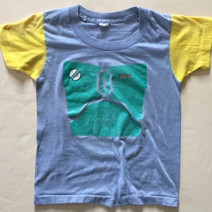 May include: A light blue t-shirt with yellow sleeves and a green and blue graphic of a Star Wars character, Boba Fett, with the text "STAR WARS" and "BOBA FETT".