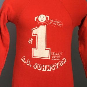 May include: Red long-sleeve sweatshirt with a white graphic of a cartoon character standing on top of the number one. The text "It's Great At The Top!" is in a speech bubble above the character. The text "Ziggy" and "A.S. Johnston" are also printed on the sweatshirt.