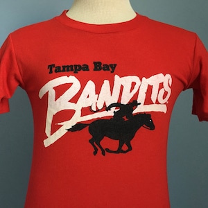 Könnte beinhalten: Rotes T-Shirt mit einer weißen Grafik eines Cowboys, der ein Pferd reitet, und dem Text "Tampa Bay Bandits".