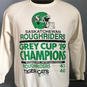 May include: White sweatshirt with green and black text and a green helmet graphic. The text reads "Saskatchewan Roughriders Grey Cup '89 Champions November 26th, 1989 Sky Dome-Toronto Roughriders 43 Tiger-Cats 40".