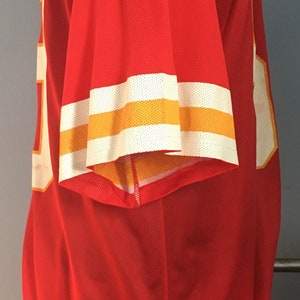May include: Red jersey with white and gold stripes on the sleeves. The jersey is made of a mesh material.