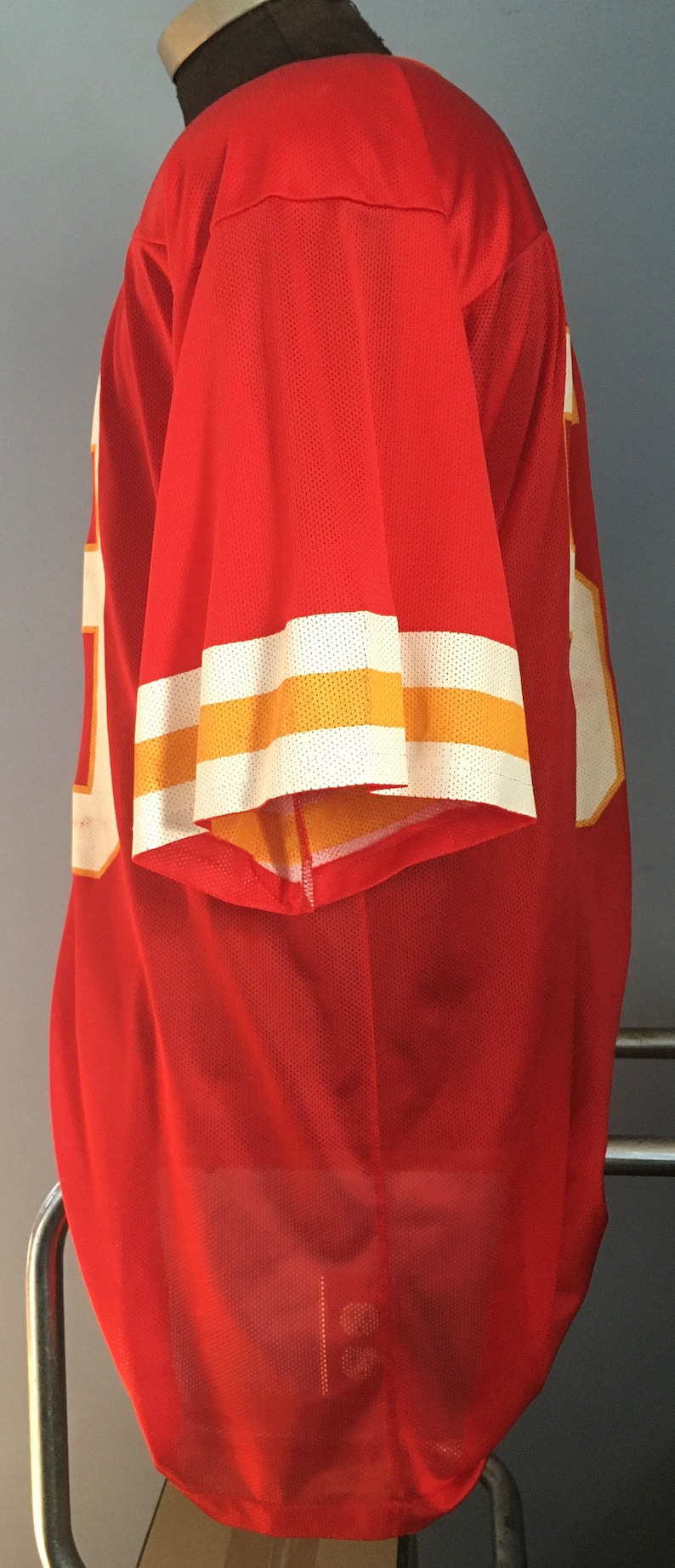 May include: Red jersey with white and gold stripes on the sleeves. The jersey is made of a mesh material.