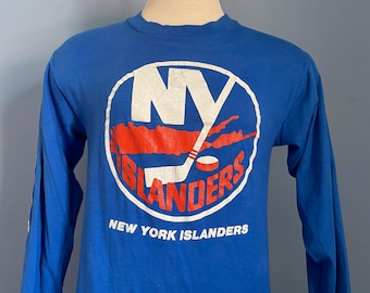 May include: A blue long-sleeve shirt featuring the New York Islanders logo. The logo is a white circle with the letters "NY" in white, a red hockey stick and puck, and the words "ISLANDERS" in red. The words "NEW YORK ISLANDERS" are printed below the logo.