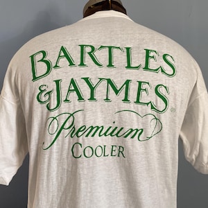 Op de afbeelding: Wit t-shirt met groene tekst die luidt "BARTLES & JAYMES Premium COOLER." De tekst is in een gestileerd lettertype. Het shirt met korte mouwen lijkt gemaakt van een zachte, ademende stof. Het shirt wordt op een mannequin getoond.
