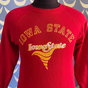 Può includere: Una felpa rossa dell'Iowa State con scritte dorate. Il testo "IOWA STATE" è arcuato nella parte superiore, con "Iowa State" in un font stilizzato sottostante. Un disegno giallo e oro è sotto il testo.