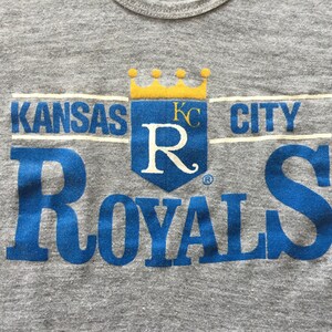 May include: A gray t-shirt with a blue and yellow Kansas City Royals logo. The logo features a crown, the letters "KC" and a large "R" with a stylized flourish. The words "Kansas City Royals" are printed in blue.