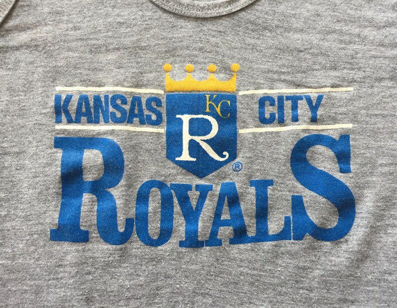 May include: A gray t-shirt with a blue and yellow Kansas City Royals logo. The logo features a crown, the letters "KC" and a large "R" with a stylized flourish. The words "Kansas City Royals" are printed in blue.