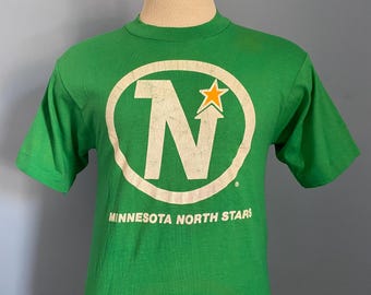 Puede incluir: Camiseta verde con el logo de los Minnesota North Stars. El logo es un círculo blanco con una "N" blanca y una estrella dorada. El texto "MINNESOTA NORTH STARS" está impreso debajo.