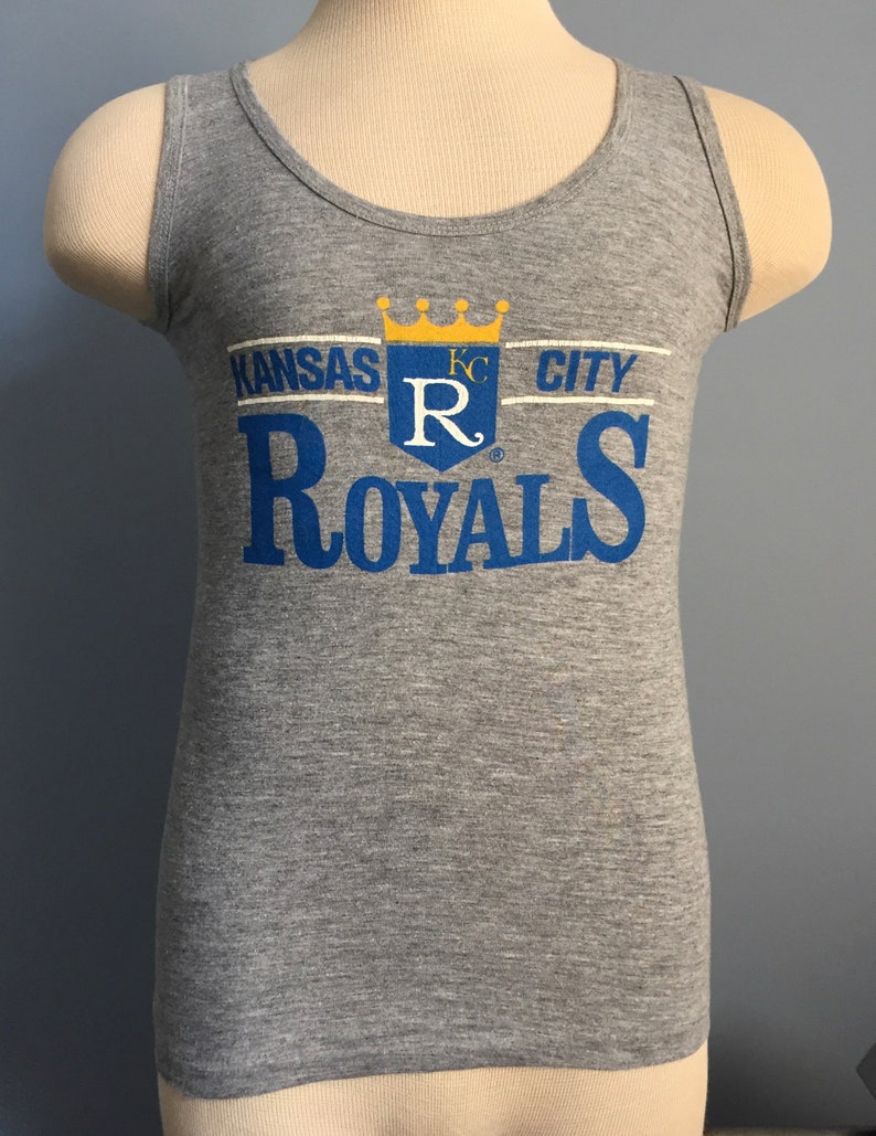 May include: Gray tank top with a blue and yellow Kansas City Royals logo. The logo features a crown, the letters "KC" and a large "R" with a stylized "ROYALS" below.