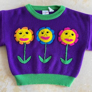 May include: A purple knitted jumper with a green collar and hem. The jumper features three embroidered flowers with yellow petals, pink lips, and green stems. A small tag is visible on the collar.