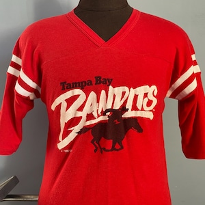 May include: Red vintage jersey-style shirt with white stripes on the sleeves. The shirt features the text "Tampa Bay Bandits" in white, along with a graphic of a rider on a horse.