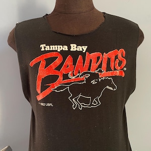 Pode incluir: Camiseta preta sem mangas com o texto "Tampa Bay Bandits" em vermelho e um gráfico de um cavalo e um cavaleiro. A camiseta também inclui o texto "1982 USFL". A gola da camiseta tem um acabamento desgastado.